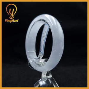 May include: A smooth, translucent white jade bangle bracelet with a polished finish. The circular bangle is displayed on a clear acrylic stand and features a small rectangular white tag with blue text attached to the inner rim of the jewelry piece.