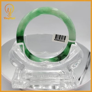 Puede incluir: Un brazalete de jade verde, exhibido en un soporte decorativo transparente. El brazalete tiene diferentes tonos de verde. Una etiqueta blanca con el texto "VDG62112" está adherida al brazalete. El brazalete tiene forma circular.