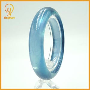 May include: A translucent blue bangle bracelet. The bracelet is a solid band of light blue material, with a smooth, polished surface. The bracelet is displayed on a clear stand against a white background. The logo "YingMart" is in the upper left corner.