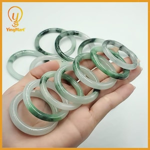 Peut inclure: Plusieurs bracelets en jade de différentes nuances de vert et de blanc. Les bracelets circulaires lisses sont présentés dans une main, mettant en valeur leurs variations de couleurs naturelles et leur qualité translucide. Bijoux populaires.