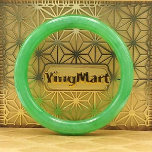 May include: A translucent, light green jade bangle bracelet. The bracelet is circular and smooth, with a polished finish. The background features a gold-colored box with a geometric pattern and the text "YingMart".