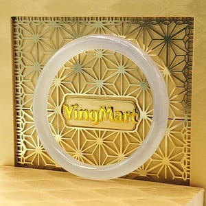 May include: A translucent jade bangle bracelet is centered in the image, set against a gold-colored background with a geometric pattern. The text "YingMart" is visible in gold lettering. The bracelet is a complete circle, showcasing its smooth, polished surface.