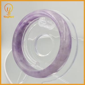 May include: A translucent, lavender-colored jade bangle bracelet. The bracelet is displayed on a clear, circular stand. The jade has a smooth, polished surface and a subtle variation in color, with lighter and darker shades of purple.