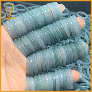 May include: A collection of pale green jade rings. The rings are stacked on fingers, showcasing their smooth, polished surfaces. The rings have a translucent quality, with subtle variations in color. The background is filled with more jade rings.