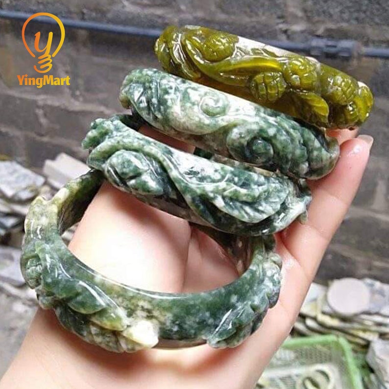 Pode incluir: Quatro pulseiras de jade verde e branco com intrincados entalhes florais. As pulseiras est&atilde;o empilhadas umas sobre as outras e est&atilde;o sendo seguradas por uma m&atilde;o.