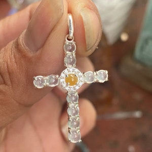 May include: A silver cross pendant featuring a central yellow gemstone encircled by small clear stones. The cross arms and stem are adorned with oval-shaped, light-colored stones. The pendant is held up by a silver loop.