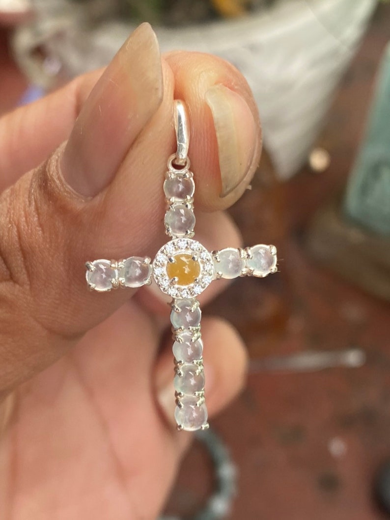 May include: A silver cross pendant featuring a central yellow gemstone encircled by small clear stones. The cross arms and stem are adorned with oval-shaped, light-colored stones. The pendant is held up by a silver loop.