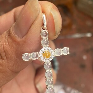 May include: A silver cross pendant with a central yellow gemstone encircled by small clear stones. The cross arms and vertical bar are adorned with clear, round gemstones. The pendant is held by a hand.