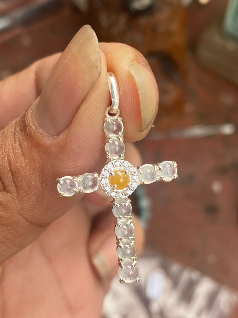 May include: A silver cross pendant with a central yellow gemstone encircled by small clear stones. The cross arms and vertical bar are adorned with clear, round gemstones. The pendant is held by a hand.