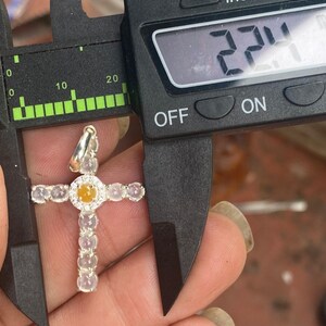 May include: A silver cross pendant with a central yellow gemstone surrounded by a halo of small diamonds. The cross is measured at 2.24 inches using a digital caliper. The cross arms are adorned with oval-shaped, light-colored gemstones.