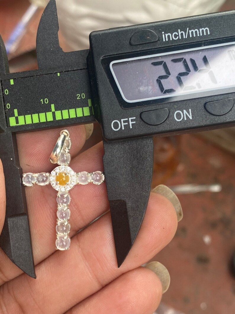 May include: A silver cross pendant with a central yellow gemstone surrounded by a halo of small diamonds. The cross is measured at 2.24 inches using a digital caliper. The cross arms are adorned with oval-shaped, light-colored gemstones.