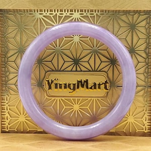 Könnte beinhalten: Ein lavendelfarbenes Jade-Armreif-Armband. Das glatte, kreisförmige Armband wird vor einem goldenen Hintergrund mit einem geometrischen Muster und dem Text "YingMart" präsentiert. Das Armband ist ein weiches, durchscheinendes Lila.