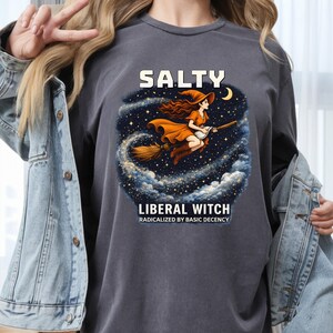 Puede incluir: Camiseta gris oscuro con la palabra "SALTY" sobre un gráfico de una bruja en una escoba. La bruja lleva un vestido y un sombrero naranja, volando por un cielo estrellado. Debajo del gráfico, el texto dice "LIBERAL WITCH RADICALIZED BY BASIC DECENCY."