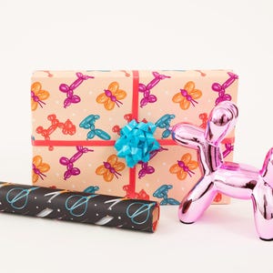 Puede incluir: Un animal de globo rosa con forma de perro, un regalo envuelto en papel con un patrón de animales de globo coloridos y un rollo de papel de regalo con un patrón blanco y negro de tijeras.
