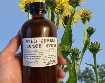 May include: A brown glass bottle labelled "Wild Cherry Cough Syrup" with a white label and black text. The label features a rainbow graphic and the word "Organic". Yellow flowers and green foliage are in the background.