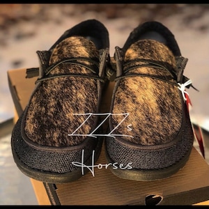 Puede incluir: Un par de zapatos negros de deslizamiento con un patrón de piel de vaca marrón y negro. Los zapatos tienen una suela de goma negra y un forro de tela negro.