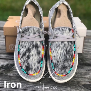 Puede incluir: Zapatillas grises de deslizamiento con un patrón de piel de vaca y un borde de cuentas de colores. Los zapatos están sobre una superficie de madera con el texto "Iron Horses" visible.