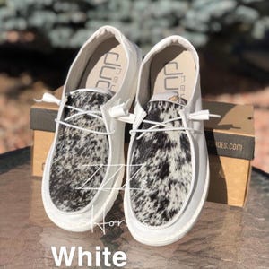 May include: A pair of white slip-on shoes with black and white cowhide-patterned vamps. The shoes have white laces and a white sole. The word "White" is written in white text at the bottom of the image.