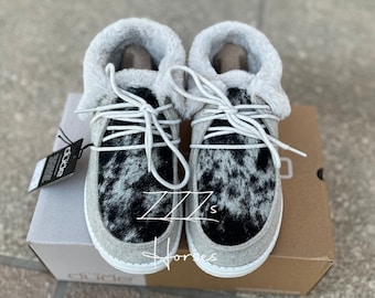 May include: A pair of Dude brand slip-on shoes with a cowhide pattern in black and white. The shoes have a gray felt upper, white laces, and a fluffy, light gray faux fur lining. The shoes are on a cardboard box.