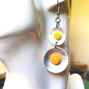Peut inclure: Boucles d'oreilles pendantes en argent avec deux disques ronds. Chaque disque a un cercle jaune au centre, ressemblant à un œuf au plat.