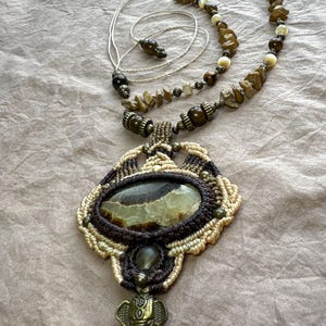 May include: A handcrafted necklace featuring a large, oval-shaped green and brown stone pendant, intricately woven with brown and beige macrame. The necklace includes a brass elephant charm and a beaded chain with natural stone accents.