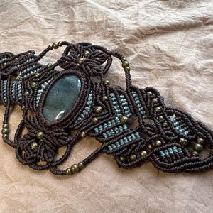 May include: A handcrafted macrame necklace featuring a central oval-shaped stone in shades of gray and green. The necklace is made with dark brown cord and accented with small brass beads. The intricate knotting creates a detailed, textured design.