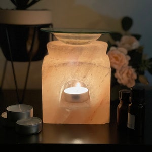 May include: A Himalayan salt lamp diffuser with a glass dish on top and a small candle burning inside. The diffuser is on a dark surface with two small candles and two bottles of essential oil.