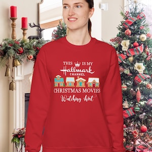 May include: A red sweatshirt with the text "THIS IS MY Hallmark CHANNEL CHRISTMAS MOVIES Watching shirt" in white. The design includes illustrations of gingerbread houses. The sweatshirt is part of a Christmas-themed collection.