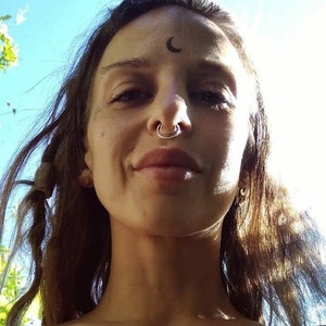 May include: A person with long brown hair, a nose ring, and a crescent moon tattoo on their forehead. The person is smiling, and the background is a bright blue sky with sunlight.