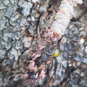 May include: A close-up of a brown stick wrapped in colourful fabric with a small owl charm and a green bead attached. The stick is resting against a rough, textured tree bark.