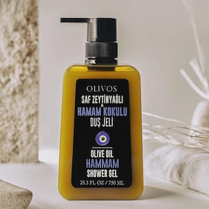 Puede incluir: Una botella cuadrada, teñida de amarillo, de gel de ducha OLIVOS Olive Oil Hammam con una etiqueta negra. La etiqueta presenta texto en turco e inglés, junto con un símbolo de ojo azul. La botella contiene 750 ml.