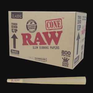 Peut inclure: Une boîte en carton brun étiquetée RAW Classic avec le texte 800 PER BOX et KING SIZE, comportant des flèches pointant vers le haut et les mots THIS END UP. Sous la boîte se trouve un cône de papier à rouler brun clair avec RAW imprimé dessus.