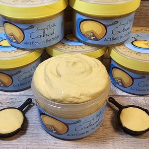 May include: A collection of yellow jars with blue lids and labels that say "Some of Y'all Cornbread Ain't Done In The Middle". The jars are filled with a yellow, creamy substance. There are also two small black cast iron skillets with yellow, creamy substance inside.