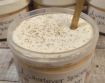 May include: A clear plastic container filled with a light brown substance topped with white whipped cream and brown sprinkles. The container has a label that reads "Whatever Spices Your Pumpkin Latte".