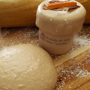 May include: A ball of dough sits on a wooden surface covered in white powder. A jar with a brown lid and the text "Bready or not, Here I crumb!" is next to the dough.