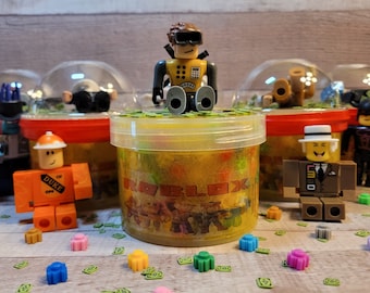 May include: A yellow and black Roblox character figurine sits on top of a clear plastic container filled with yellow slime. The container has a red and white label that says "Roblox". There are other Roblox figurines and containers in the background.