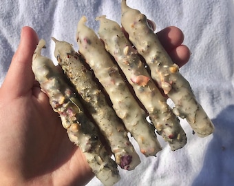 May include: A set of six white candles with dried flowers and herbs embedded in the wax. The candles are arranged in a hand, with the tips of the candles pointing towards the right.