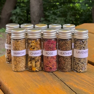 May include: Ten small glass jars with silver lids filled with dried herbs and flowers. The jars are labeled with the names of the herbs: Hyssop, Yarrow, Chamomile, Rose, Heather, Lavender.