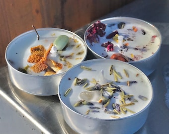 May include: Three small, round candles in silver containers. Each candle has a white wax base with decorative elements. One candle has a green stone and orange peel, another has red petals and a brown stone, and the third has lavender and a clear stone.