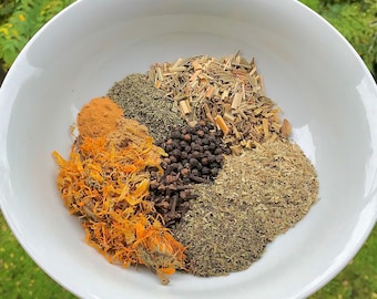 May include: A white bowl filled with various dried herbs and spices, including lemongrass, cinnamon, cloves, black peppercorns, and dried flowers.