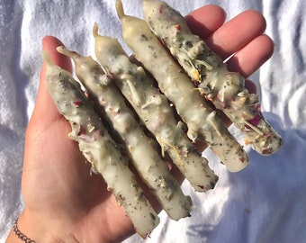 May include: A set of five white candles with dried herbs and flowers embedded in the wax. The candles are arranged in a hand, with the tips pointing towards the camera.