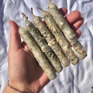May include: Five white candles with dried herbs and flowers embedded in the wax. The candles are arranged in a hand.