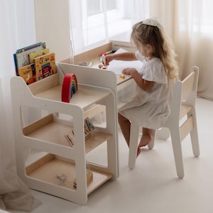 Op de afbeelding: Een witte houten kinderbureau- en stoelset. Het bureau heeft een ingebouwde boekenplank met kleurrijke boeken en een houten regenboog speelgoed. Een kind zit aan het bureau en tekent op een vel papier.