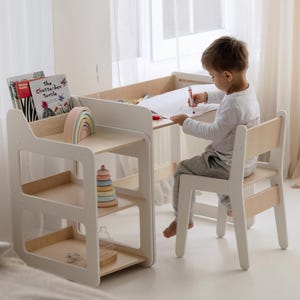 Op de afbeelding: Een witte en natuurlijke houten kinderbureau en stoel set. Het bureau heeft een ingebouwd schap en een aparte stellingkast met drie planken. Een kind zit aan het bureau en tekent met een rode stift. Boeken en een houten regenboog staan op de planken.