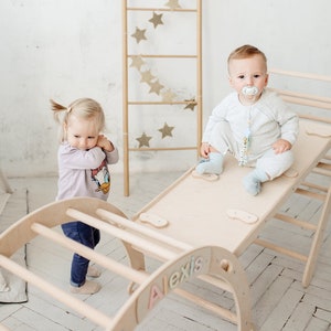 May include: Two toddlers playing on a wooden climbing structure. The structure features a climbing wall, a slide, and a flat platform. The platform has the name "Alexis" written on it.