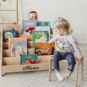 May include: A wooden bookshelf with a personalized nameplate that reads "Alexis". The bookshelf is filled with colorful children's books. Two children are interacting with the bookshelf, one is looking at the books and the other is holding a book.
