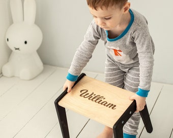Pode incluir: Um banquinho de madeira com pernas pretas e o nome "William" gravado na parte superior. Uma criança pequena está segurando o banquinho.
