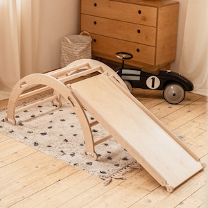 May include: A wooden climbing frame with a slide attached. The frame is made of natural wood and has a ladder on one side. The slide is made of a smooth, light-colored wood and is attached to the frame with metal brackets. The frame is sitting on a rug with a white background and black dots. There is a toy car in the background.