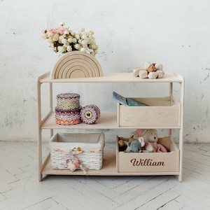 May include: A wooden shelf unit with three shelves, featuring a white wicker basket, a rainbow wooden toy, and a wooden box labeled "William".