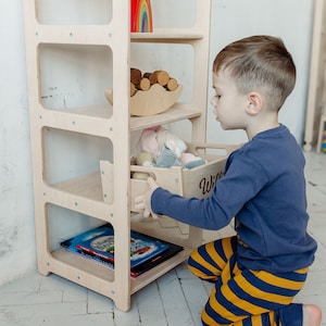 May include: A light wood bookshelf with four shelves and a drawer. The drawer is labeled "Willie". A child is kneeling and opening the drawer. The bookshelf is filled with toys, books, and other items.